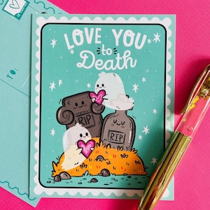 May include: A greeting card with the text "LOVE YOU to Death" on a teal background. The card features cartoon ghosts holding pink hearts near tombstones. A teal envelope and a gold pen are in the background.