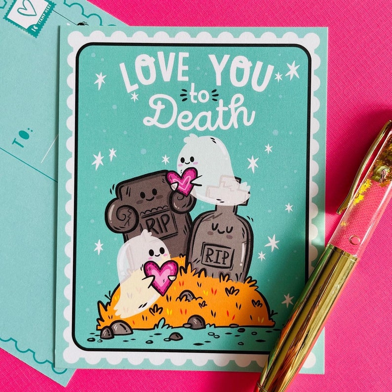 May include: A greeting card with the text "LOVE YOU to Death" on a teal background. The card features cartoon ghosts holding pink hearts near tombstones. A teal envelope and a gold pen are in the background.