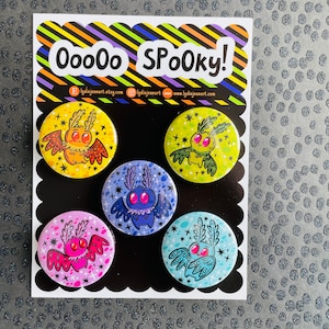 May include: Five colorful round buttons with a spooky moth design. Each button features a different color background and a moth with large eyes and wings. The text "Ooooo Spooky!" is at the top of the card.