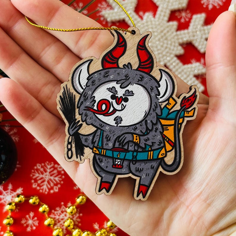 May include: A wooden Christmas ornament shaped like a gray, furry monster with red horns, a red tongue, and a blue and yellow backpack. The monster is holding a black chain in its right hand.