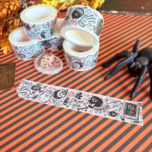 May include: A collection of Halloween-themed washi tape rolls with black and white illustrations of ghosts, bats, and other spooky elements. A decorative strip of tape with the words "Creep It Real" is also visible.
