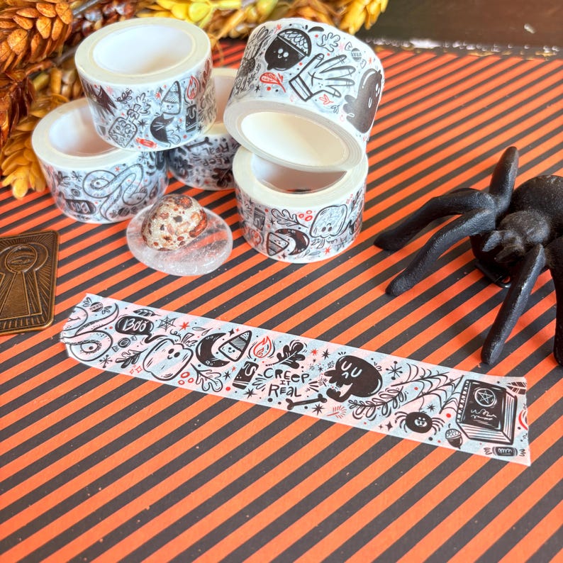 May include: A collection of Halloween-themed washi tape rolls with black and white illustrations of ghosts, bats, and other spooky elements. A decorative strip of tape with the words "Creep It Real" is also visible.