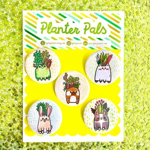 May include: Five colorful round buttons with a white background and a yellow border. Each button features a different animal with plants growing on its head. The animals are a frog, a sloth, a bunny, a cat, and a dog. The buttons are labeled "Planter Pals" and include the Etsy shop name and website.