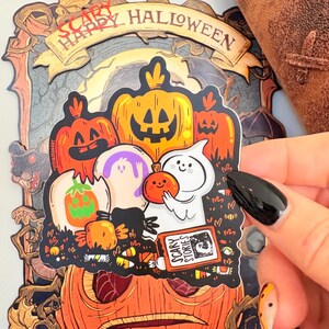 May include: A Halloween-themed sticker featuring cartoon pumpkins, ghosts, and candy. The sticker has the words "Scary Happy Halloween" at the top and "Scary Stories" on a book. The color palette includes orange, purple, white, and black.