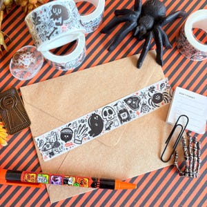 May include: A Halloween-themed flat lay featuring decorative washi tape rolls with skull and ghost designs, a brown envelope with matching tape, a black spider, and a pen. The background is orange and black striped.