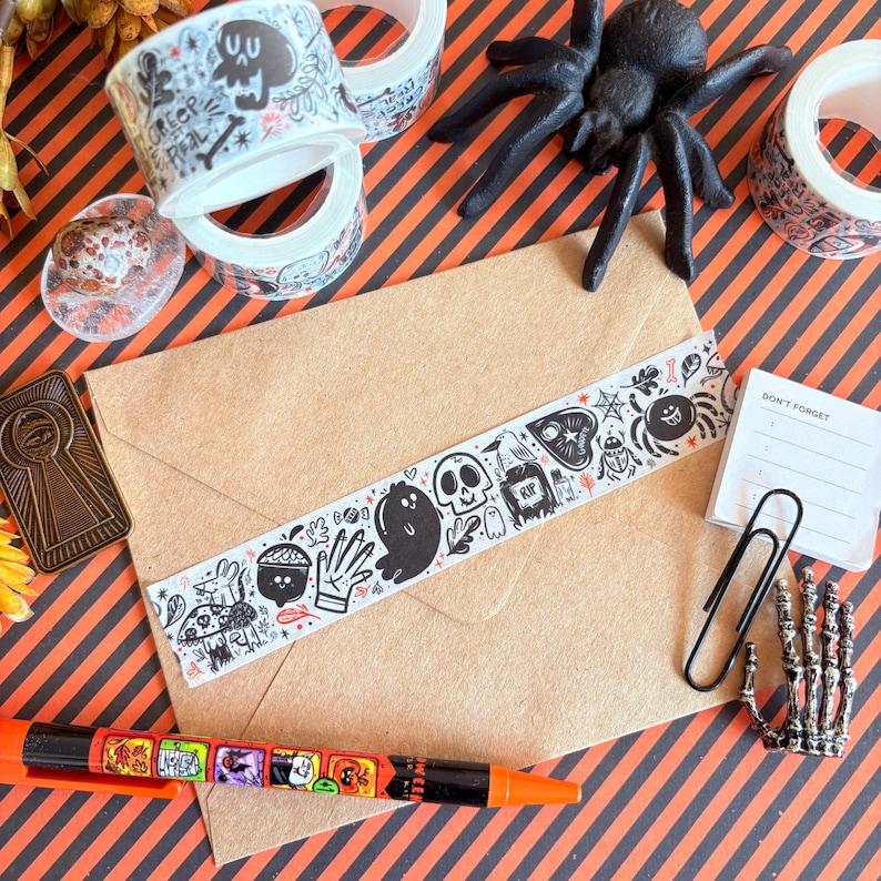 May include: A Halloween-themed flat lay featuring decorative washi tape rolls with skull and ghost designs, a brown envelope with matching tape, a black spider, and a pen. The background is orange and black striped.
