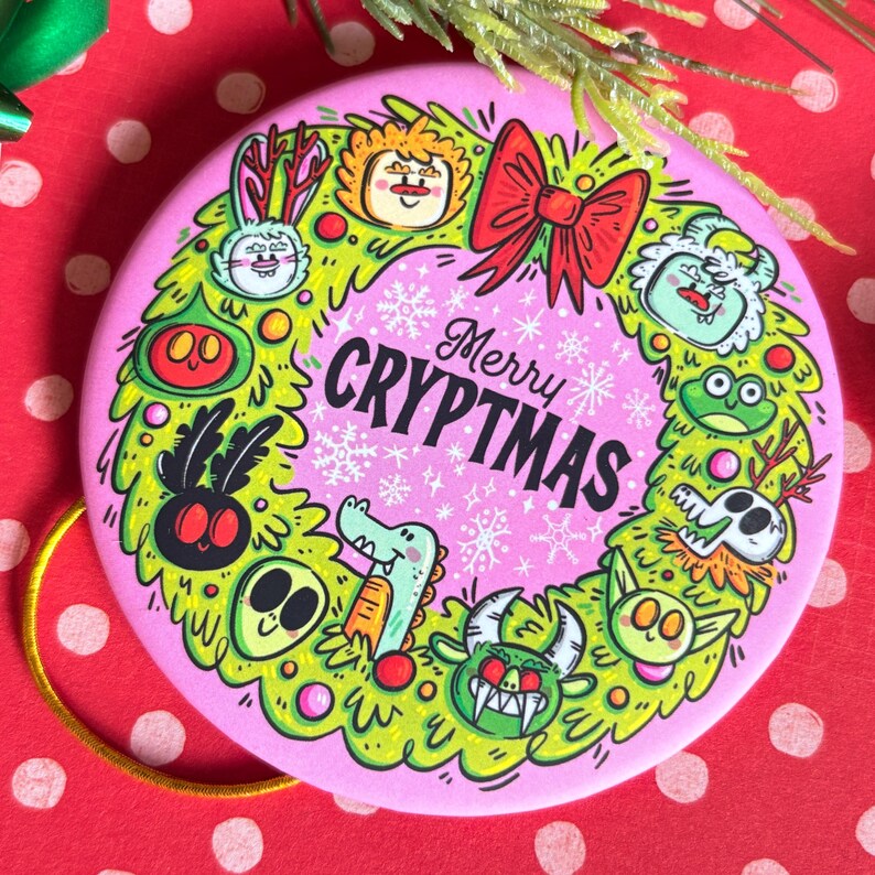 Puede incluir: Un adorno navide&ntilde;o redondo rosa con un dise&ntilde;o de corona verde. La corona presenta monstruos de dibujos animados y un lazo rojo. El texto "Merry CRYPTMAS" est&aacute; en el centro. El adorno est&aacute; sobre una superficie roja con lunares blancos.