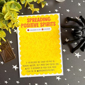 Puede incluir: Una pegatina amarilla con el texto "SPREADING POSITIVE SPIRITS" en rojo. La pegatina tiene un borde festoneado y un patr&oacute;n de figuras fantasmales. La pegatina tambi&eacute;n incluye informaci&oacute;n de redes sociales e instrucciones para compartir vibraciones positivas.