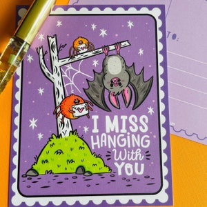 May include: A purple postcard with a cartoon bat hanging upside down and two spiders. The text "I MISS HANGING With YOU" is written in white. The postcard has a white scalloped border and is decorated with stars.