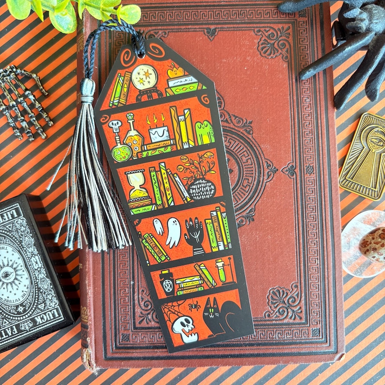 以下が含まれることがあります： 黒いタッセルが付いたハロウィーンをテーマにしたブックマーク。薬瓶、本、骸骨が描かれた本棚のデザインです。ブックマークはオレンジと黒で、装飾的なボーダーと黒い紐が付いています。