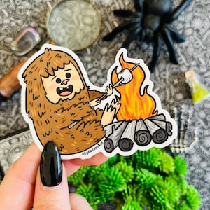 May include: A brown furry creature with a big smile roasts a marshmallow over a campfire. The creature is holding a stick with a marshmallow on the end. The campfire is made of logs and has flames coming from it. The background is a white sticker with the text "LydiaJeanArt" in the bottom right corner.