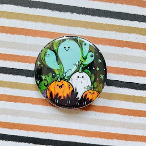 May include: A round button with a Halloween theme. It features a green background with three white ghosts, two orange pumpkins with black eyes and mouths, and a black and white ghost with a star pattern. The button has a black border.