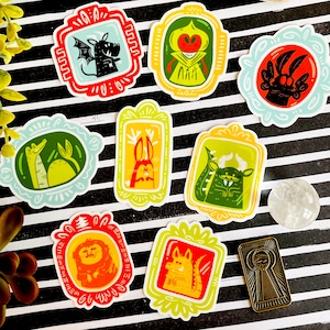 May include: Nine colorful stickers featuring various cartoon monsters in ornate frames. The stickers are on a black and white striped background.