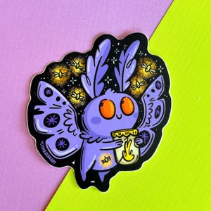 May include: A die-cut sticker featuring a cartoon purple moth with large orange eyes and patterned wings. The moth holds a glowing yellow jar, surrounded by small fireflies against a black background. The text LYDIAJEANART is printed on the side.