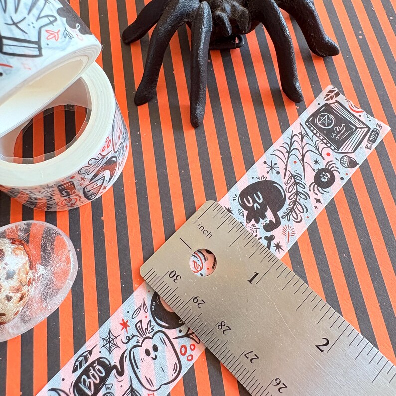 May include: Halloween-themed washi tape and ruler with a black plastic spider. The tape features a light blue background with spooky illustrations, including skulls, pumpkins, and the word "Boo". The ruler measures in inches.