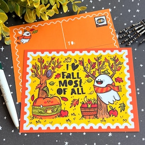 Könnte beinhalten: Orangefarbene Postkarte mit gelbem Rand und herbstlicher Illustration. Das Design zeigt einen Geist, einen Kürbis, Bäume und den Text "I Fall Most of All". Ein orangefarbener Umschlag mit Vogelgrafik befindet sich hinter der Postkarte.