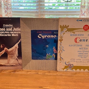 May include: Three vintage vinyl record albums. The album covers feature artwork and text. One album is titled "Romeo and Julie" with a ballet scene. Another is "Cyrano". The third is "Camelot" with illustrations and text.