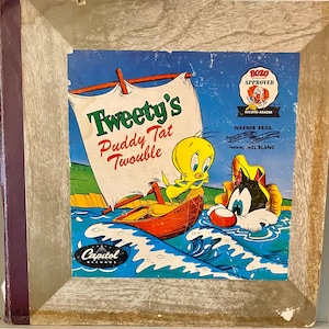 Puede incluir: Una portada de álbum de vinilo vintage con Tweety Bird y Silvestre el gato. El álbum se titula "Tweety's Puddy Tat Twouble" y presenta el logotipo de Warner Bros. y el nombre Mel Blanc. La portada del álbum está en un marco de madera.