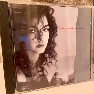May include: A CD case featuring a black and white image of a woman with curly hair. The text "gloria estefan cuts both ways" is in red. The CD case is in a clear plastic case.