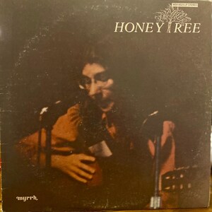 Puede incluir: Funda de álbum de vinilo vintage para "Honey Tree" con fondo marrón oscuro. La portada del álbum presenta una imagen borrosa de una persona tocando la guitarra y cantando a un micrófono. El título del álbum y el nombre del artista están en texto blanco.