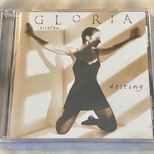 May include: CD case for the album "Destiny" by Gloria Estefan. The cover features a woman in a black dress kneeling with arms outstretched. The album title is in cursive, and the artist's name is at the top.