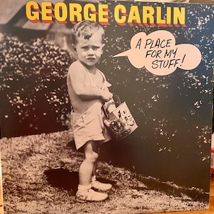 Puede incluir: Una portada de álbum en blanco y negro con el texto "GEORGE CARLIN" en amarillo y rojo. Un niño pequeño sostiene un cubo con el texto "A PLACE FOR MY STUFF!" en un bocadillo.