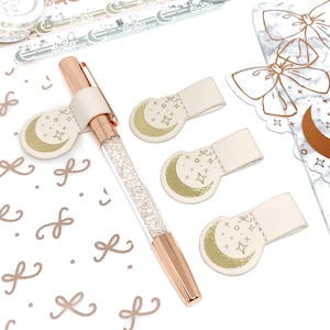 May include: Four beige leather pen holders with a gold crescent moon and stars design. The holders are on a white surface with a pen, washi tape, and stickers with a similar design.