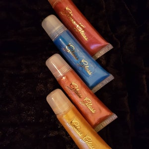 May include: Four tubes of lip gloss in shades of red, blue, copper, and gold. Each tube has a clear applicator and a gold script logo that reads "Glass Gloss". The tubes are arranged diagonally on a dark, textured surface.