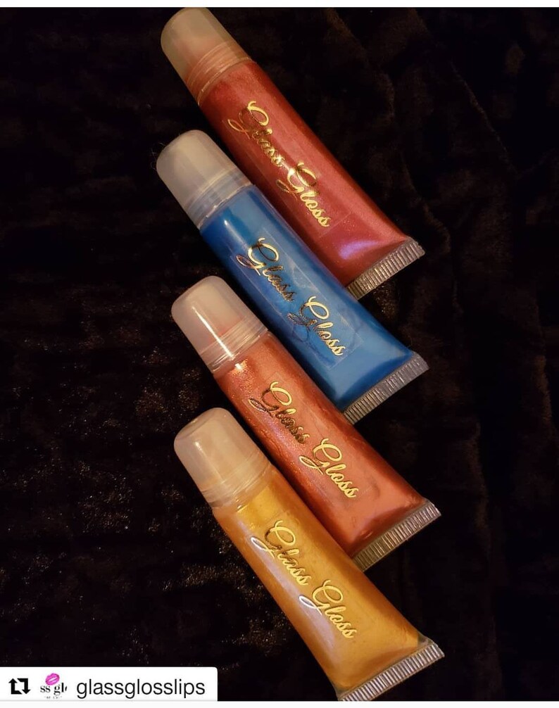 May include: Four tubes of lip gloss in shades of red, blue, copper, and gold. Each tube has a clear applicator and a gold script logo that reads "Glass Gloss". The tubes are arranged diagonally on a dark, textured surface.