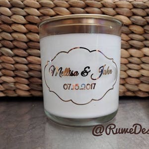 May include: A white candle in a glass jar with a gold-colored lid. The candle has a decorative label with the names "Melissa & John" and the date "07.15.2017". The background features a woven basket.