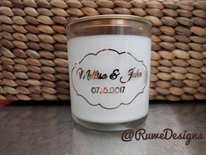 May include: A white candle in a glass jar with a gold-colored lid. The candle has a decorative label with the names "Melissa & John" and the date "07.15.2017". The background features a woven basket.