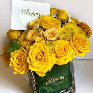 May include: A vibrant floral arrangement featuring bright yellow roses and other flowers in a clear glass vase. A white card with the word "Blooms" in gold lettering is placed in the arrangement. The vase has a green interior with the word "Blooms" in gold lettering.