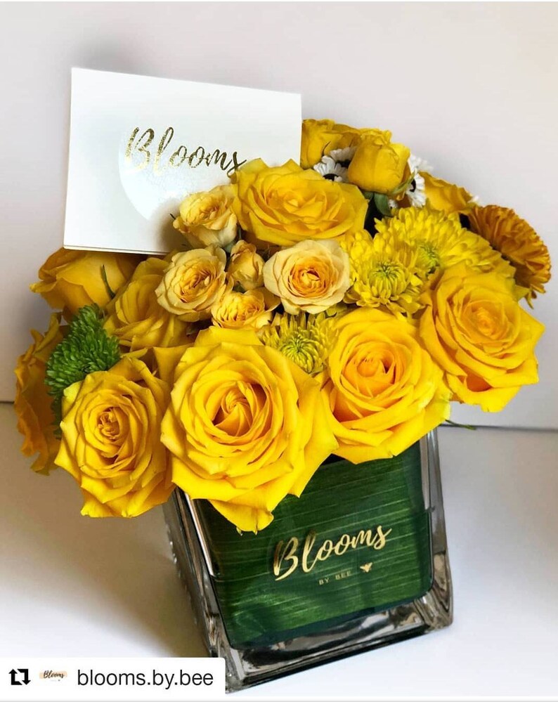 May include: A vibrant floral arrangement featuring bright yellow roses and other flowers in a clear glass vase. A white card with the word "Blooms" in gold lettering is placed in the arrangement. The vase has a green interior with the word "Blooms" in gold lettering.