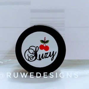 May include: A black jar with a white label that says "Suzy" in a cursive font. The label also features two red cherries with green stems and leaves.
