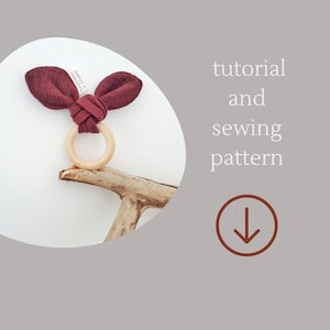May include: A tutorial and sewing pattern for a burgundy fabric baby teether with wooden ring. The teether is tied with a knot and has two bunny ear-shaped fabric pieces. The image includes the text "tutorial and sewing pattern" and a downward arrow.