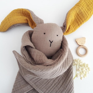 May include: A soft bunny toy with mustard and grey ears, a brown head, and a matching scarf. A wooden bear-shaped teether and a small bouquet of white flowers are also present.