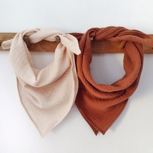 May include: Two cotton bandana scarves hanging on a wooden rack. One scarf is a light beige colour and the other is a burnt orange colour.