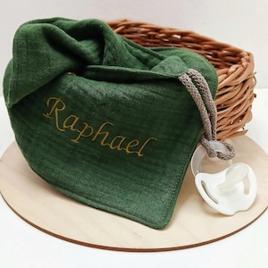 May include: A dark green baby bib with the name "Raphael" embroidered in gold thread. A white pacifier with a clear ring is attached. The bib is on a wooden round surface, with a wicker basket in the background.