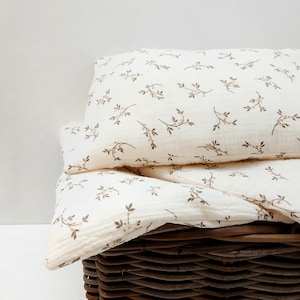 Puede incluir: Un juego de almohada y manta de color crema con un delicado estampado floral marrón, dentro de una cesta de mimbre marrón tejida. La ropa de cama tiene un aspecto suave y texturizado, lo que sugiere comodidad y una estética natural. La cesta añade un toque rústico.