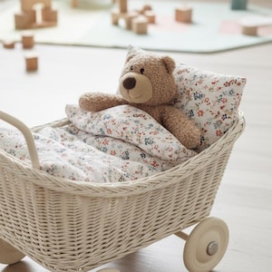 Puede incluir: Un cochecito de muñecas de mimbre color crema con un oso de peluche marrón, ropa de cama floral y una almohada. El cochecito tiene cuatro ruedas y un asa. La escena está ambientada en una habitación luminosa con juguetes de madera.