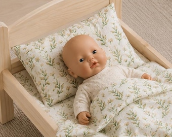 May include: A light-colored wooden doll bed featuring a mattress, pillow, and blanket with a green and white floral pattern. A small doll with a smooth, light-toned face and a long-sleeved cream-colored top lies tucked under the floral blanket.