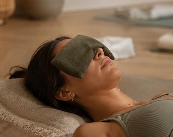 May include: A sage green rectangular eye pillow resting over the eyes of a person lying on a yoga mat. The person wears a matching sage green ribbed sports bra and is positioned on a light-colored pillow, with a small dark glass bottle nearby.