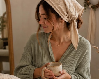 May include: A sage green long-sleeved top, a beige headscarf, a crescent-shaped beige pillow, a small sage green drawstring pouch, an open book, a small beige bear-shaped security blanket, and a sage green bolster pillow.