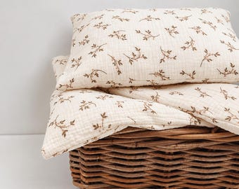 May include: A cream-coloured pillow and folded blanket set featuring a repeating brown botanical pattern of small leaves and berries. The fabric has a visible crinkled texture, and the items are resting inside a woven brown wicker basket.