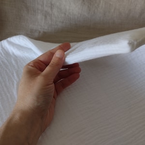 May include: White cotton muslin fabric with a textured weave. The fabric is being held in a hand, showing its soft and drapey texture.