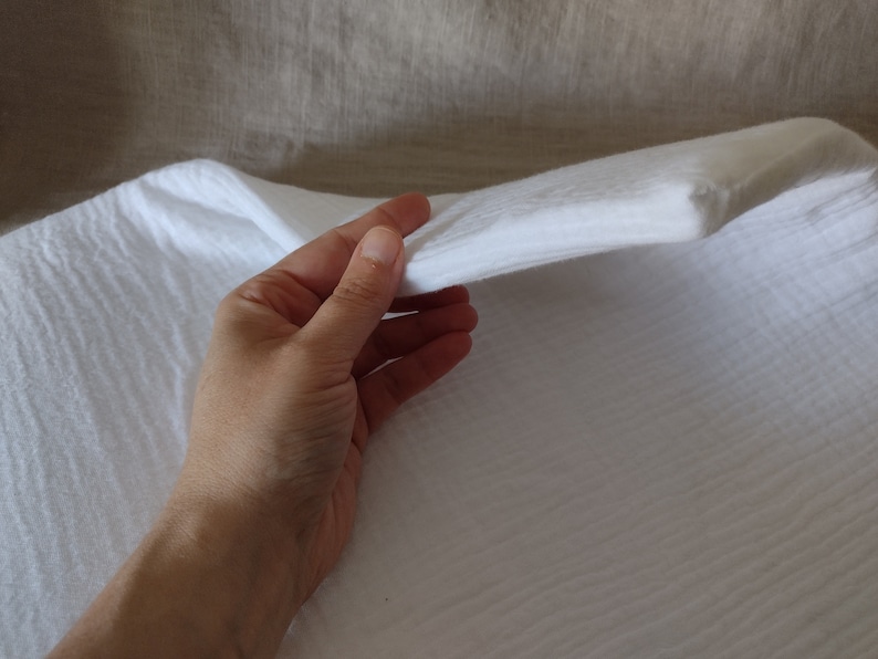 May include: White cotton muslin fabric with a textured weave. The fabric is being held in a hand, showing its soft and drapey texture.