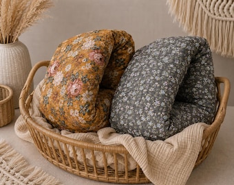 May include: Two folded fabric cushions inside a woven wicker basket. One cushion features a mustard yellow background with a floral pattern of pink roses and white daisies. The other cushion is dark gray with a small white and blue floral print.