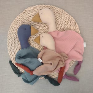 May include: Three plush goose toys in blue, pink, and beige, each with a yellow beak and a knotted tail. The geese are arranged on a woven circular mat, creating a soft and playful aesthetic.
