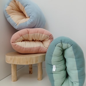 May include: Three soft, folded, rectangular fabric cushions made of crinkled muslin material. One cushion is light blue and cream, one is dusty pink and cream, and one is sage green and light green, featuring a small white PureCotton Studio tag.