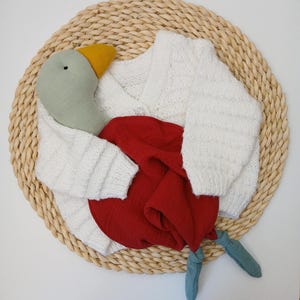 May include: A white knitted cardigan with a button closure, paired with a red fabric and a stuffed goose toy. The goose has a green head, yellow beak, and blue feet. The items are arranged on a woven, circular mat.